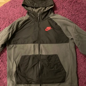 Nike hoodiejackor svart & grå L - Två snygga Nike hoodiejackor i storlek L. Ena jackan är svart med vit Nike-logga och reflexdetalj, den andra är grå och svart med röd logga. Båda har dragkedja, huva och stora fickor framtill. Perfekta för chill eller träning.