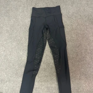 Svarta ridleggings från Equestrian Stockholm - Snygga svarta ridleggings med hög midja och silikonmönster för extra grepp. Stretchigt material som sitter tight och bekvämt. Perfekta för ridning och träning, med diskret logga på benet och praktiska fickor på sidorna. De har ett lite litet hål som jag har sytt men de syns kanppast!