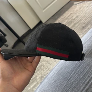 Svart Gucci keps med GG-mönster - Snygg svart keps från Gucci med klassiskt GG-monogram i tyget och ikoniska gröna och röda ränder på sidan. Justerbar rem bak i läder med guldfärgad spänne. Perfekt accessoar för dig som vill sticka ut med lyxig streetstyle. Har själv tagit bort lappen👍