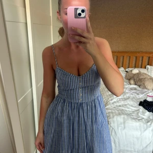 Blåvit randig klänning från Abercrombie & Fitch - Säljer en söt blåvit randig klänning från Abercrombie & Fitch i storlek S. Klänningen har smala axelband, knappar framtill och en smockad rygg för extra komfort. Perfekt för varma dagar och har en luftig passform.