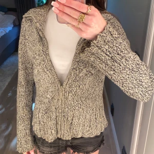 Beige och svart stickad kofta från LinDEX - Säljer en mysig stickad kofta från LinDEX i beige och svart melerad färg. Koftan har dragkedja framtill, lång ärm och bred krage. Perfekt att slänga på sig när det är kyligt ute.