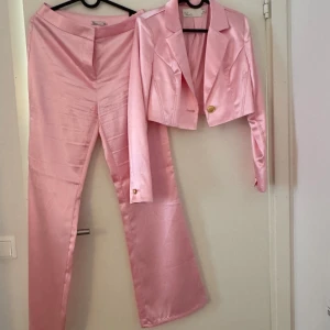 Rosa satin kostym från NLY Trend - Snygg tvådelad kostym i glansigt rosa satin från NLY Trend. Kavajen är croppad med guldknapp och klassisk krage, byxorna är raka och höga i midjan. Aldrig använt.
