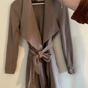 Beige trenchcoat från ONLY med bälte - Snygg beige trenchcoat från ONLY i storlek XS. Jackan har långa ärmar, bred krage och knyts i midjan med ett bälte för en clean och stilren look. Perfekt för lager-på-lager och passar till många outfits.