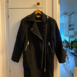 Svart kappa med dragkedjor  - Svart bikerkappa med coola silvriga dragkedjor och stora fickor. Jackan har en rak passform, bred krage och långa ärmar. Perfekt för dig som gillar en stilren look. Materialet känns som ullblandning och passar grymt till höst, vinter och vår.