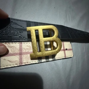 Burberry bälte med guldspänne - Snyggt bälte från Burberry med klassiskt rutigt mönster i beige, svart och rött. Bältet har ett stort guldigt spänne med initialerna 'B'. Perfekt accessoar för att lyfta din outfit.