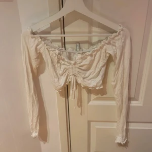 Vit croppad blus från NLY Trend - Säljer en vit croppad blus från NLY Trend med långa ärmar och resår vid axlar och ärmslut. Toppen har rynkade detaljer framtill och en söt volangkant längs halsringningen och nederkanten.