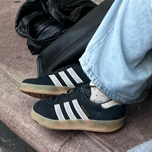 Adidas Gazelle svarta mocka 37 - Svarta Adidas Gazelle sneakers i mocka med klassiska vita ränder och gummisula. Snygg retrostil med guldfärgad logga på sidan och stickade detaljer vid hälen. Perfekta för dig som gillar streetwear och tidlös design.