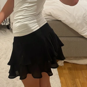 Svart volangkjol med resårmidja - Supersöt svart kjol med volanger och resår i midjan. Resåren i midjan går att vika ner så får man kjolen mer lågmidjad. Materialet känns lätt och luftigt. Storlek XS men passar bra till S och M.