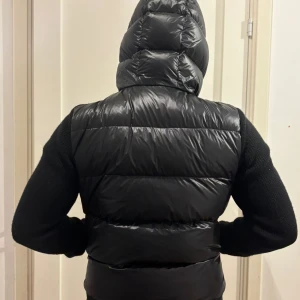 Svart hybridväst med huva från Moncler - Säljer en svart väst från Moncler med stickade ärmar och quiltad kropp. Västen har dragkedja framtill, två fickor med dragkedja och en vadderad huva. Perfekt för kyliga dagar och har en sportig look. Är 181 så du ser hur de sitter på mig ! Säljs pga den börjar bli för liten !