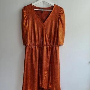 Glittrig kopparfärgad omlottklänning M - Säljer en glittrig kopparfärgad omlottklänning från Rainbow med v-ringning och trekvartsärm. Klänningen har resår i midjan och ett lätt glansigt material som ger en festlig känsla. Fint skick!