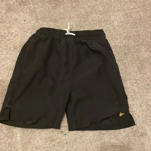 Svarta badshorts från Lyle & Scott Junior - Svarta badshorts från Lyle & Scott Junior med vit snörning i midjan och innerbyxa i mesh. Klassisk design med elastisk midja och liten gul logga på benet. Perfekta för stranden eller poolen.