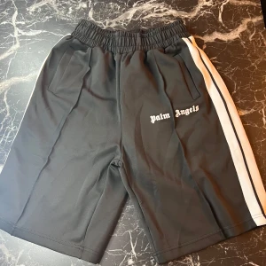 Svarta shorts från Palm Angels med vit sidoremsa - Svarta shorts från Palm Angels med vit sidoremsa längs sidorna och logotyp på vänster ben. Shortsen har elastisk midja med snörning och två sidofickor. Perfekta för en sportig och trendig look.
