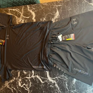 Svarta träningsshorts från Under Armour - Säljer ett par svarta träningsshorts från Under Armour med logga i färg på vänster ben. Shortsen har snörning i midjan och är tillverkade i ett lätt och snabbtorkande syntetmaterial. Perfekta för löpning eller gym.