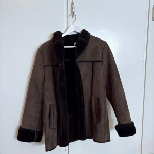 Brun pälsjacka från IsoLina - Snygg brun pälsjacka. Jackan har mjukt foder, uppvikta ärmslut och markerade sömmar. Perfekt för kalla dagar när du vill vara både varm och stilren. Klassisk rak passform och två fickor framtill.