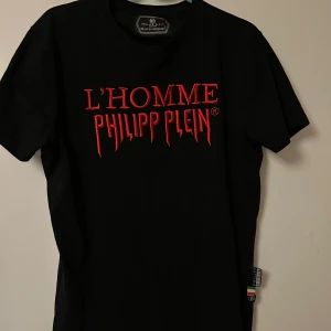 Svart Philipp Plein t-shirt med röd text - Svart t-shirt från Philipp Plein med fet röd broderad text 'L'HOMME PHILIPP PLEIN' på bröstet. Klassisk passform, korta ärmar och rund hals. Snygg detalj med logga och italiensk flagga på sidan. Perfekt för dig som vill sticka ut med en exklusiv streetstyle.