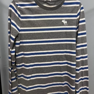 Randig långärmad tröja Abercrombie - Snygg långärmad tröja från Abercrombie & Fitch i grått med blå och vita ränder. Klassisk rund halsringning och broderad älg-logga på bröstet. Skön passform och perfekt för dig som gillar en avslappnad men stilren look.