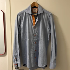 Ljusblå skjorta från Chadburry - Snygg ljusblå skjorta från Chadburry i slim fit-modell. Skjortan har orange detaljer på insidan och orange knappar, samt klassisk krage och långa ärmar. Tillverkad i 100% bomull för en skön känsla.