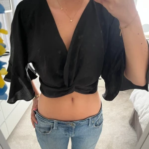 Svart omlottopp med vida ärmar - Säljer en svart omlottopp med djup v-ringning och knutdetalj framtill. Toppen har vida trekvartsärmar och är croppad, vilket ger en trendig look. Perfekt att styla med jeans eller kjol.