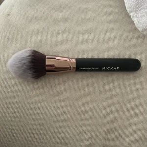 Hickap 111 Powder Deluxe sminkborste - Stor och fluffig puderborste från Hickap, modell 111 Powder Deluxe. Borsten har täta, mjuka syntetiska strån i vitt och brunt, ett roséguldfärgat fäste och svart handtag. Perfekt för applicering av löspuder eller kompaktpuder.