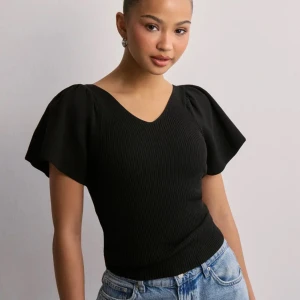 Svart ribbad topp med puffärmar och rosett - Snygg svart ribbad topp med v-ringning och korta puffärmar. Toppen har en fin rosettdetalj i ryggen och är figurnära i modellen. Perfekt att styla med jeans eller kjol för en trendig look.