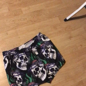 Svarta shorts med dödskallemönster - Coola svarta shorts med stora dödskallar och gröna samt lila detaljer. Perfekta för dig som vill sticka ut med ett unikt mönster. Resår i midjan för skön passform.