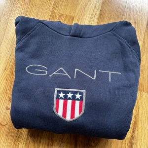 Marinblå hoodie från GANT med logga - Snygg marinblå hoodie från GANT med broderad logga och sköld på bröstet. Tröjan har huva, känguruficka och långa ärmar. Perfekt för dig som gillar klassisk och sportig stil. Materialet är mjuk bomull som känns skönt mot huden.