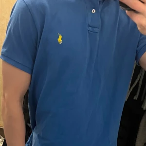 Blå pikétröja från Polo Ralph Lauren - Klassisk blå pikétröja från Polo Ralph Lauren med gul broderad logga på bröstet. Tröjan har krage, två knappar och korta ärmar. Tillverkad i bomull och har en normal passform. Perfekt för en clean och stilren look.