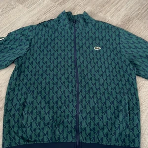 Grön mönstrad tracksuit från Lacoste - Säljer en grön mönstrad tracksuit från Lacoste med dragkedja och långärmad design. Tracksuiten har ett diskret mönster och en liten logotyp på bröstet. Perfekt för en sportig look!