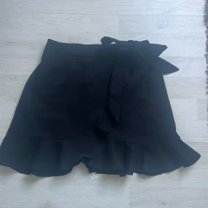Svart kjol med volang och knytband - Snygg svart kjol med volangdetaljer och ett knytband i midjan. Perfekt för att ge en elegant touch till din outfit. Kjolen har en kort längd och är lätt att matcha med olika toppar.