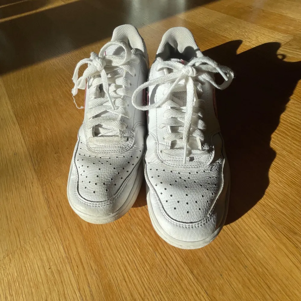 Säljer ett par vita Nike sneakers med rosa detaljer och snörning. Skorna har en klassisk design med perforeringar på tån för ventilation. Perfekta för en avslappnad stil.. Kengät.