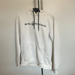 Vit hoodie från Sail Racing - Säljer en stilren vit hoodie från Sail Racing med svart text på framsidan. Den har en klassisk känguruficka och justerbar huva med svarta snören. Perfekt för en avslappnad stil.