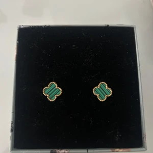 Gröna fyrklöverformade örhängen - Säljer ett par eleganta örhängen i form av fyrklöver med gröna stenar och en fin guldfärgad kant. Perfekta för att ge en touch av lyx till din outfit.