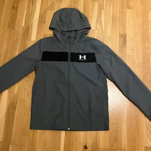 Grå vindjacka från Under Armour - Snygg grå vindjacka från Under Armour med svart panel och logga på bröstet. Jackan har en praktisk huva och dragkedja framtill. Perfekt för blåsiga dagar.