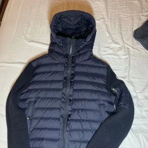 Mörkblå cardigan från Moncler - Säljer en snygg mörkblå dunjacka från Moncler med stickade ärmar och huva. Jackan har dragkedja och en liten ficka med dragkedja på ärmen. Om ni vill se mer bilder så kan ni fråga.