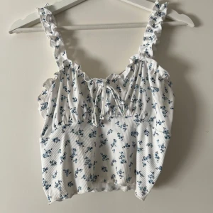 Vit blommig topp - Söt vit topp med blå blommor och volangdetaljer. Toppen har en ribbad struktur och  perfekt till sommaren! Enbart testad så i nyskick. Pris går att diskutera! 
