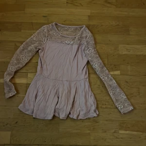 Ljusrosa klänning med spetsdetaljer - Säljer en söt ljusrosa blus med långa spetsärmar och volangkjol. Perfekt för speciella tillfällen. Den har en elegant design med spetsdetaljer som ger en romantisk känsla. Passar för åldrar 11-12