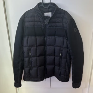Moncler jacka  - Snygg svart dunjacka från Moncler med quiltad design och dragkedja framtill. Jackan har en stilren look med Moncler-logga på ärmen och praktiska fickor med dragkedja. Perfekt för vår/höst.