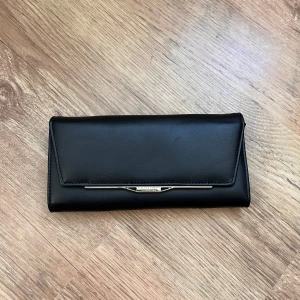 Ny och oanvänd svart plånbok med metallspänne - Elegant svart plånbok med ett stilrent metallspänne framtill. Inuti finns flera kortfack och ett fönster för ID-kort. Perfekt för att hålla ordning på dina kort och kontanter. Den är ny, dvs. helt oanvänd. 