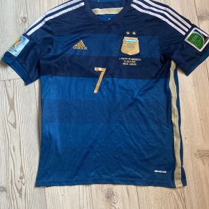 Di Maria  vm 2014 tröja - Säljer en blå fotbollströja från Adidas med guldiga detaljer och nummer 7. Tröjan har korta ärmar och tre vita ränder på axlarna. Den har även ett tryck med 'DI MARIA' på ryggen. Perfekt för fotbollsfans!