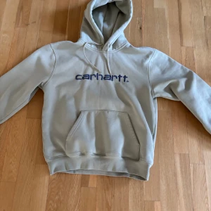 Carhartt hoodie - Säljer en beige hoodie från Carhartt med en stor ficka framtill och justerbar huva. Tröjan har långa ärmar och Carhartt-loggan tryckt på bröstet i blått. Perfekt för en avslappnad stil.