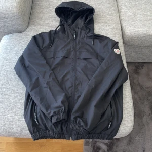 Svart vindjacka från Moncler - Säljer en stilren svart vindjacka från Moncler med huva och dragkedja. Jackan har elastiska muddar och en diskret logga på ärmen. Perfekt för blåsiga dagar. Pris kan diskuteras!!