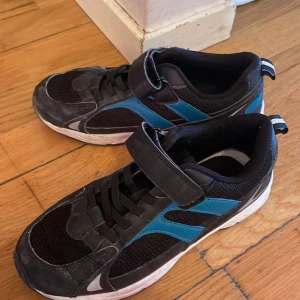 Svarta och blå sneakers med kardborreband - Säljer ett par svarta sneakers med blå detaljer och kardborreband. Skorna har en vit sula och är perfekta för vardagsbruk. De har en rund tå och är gjorda av syntetmaterial.