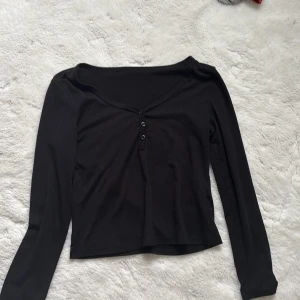 Svart långärmad topp med knappar - Säljer en stilren svart långärmad topp med knappar framtill. Perfekt för en enkel och snygg look. Toppen har en v-ringad design och är tillverkad i ett mjukt material.