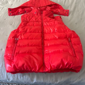 Röd dunväst från Pepe Jeans - Säljer en snygg röd dunväst från Pepe Jeans med dragkedja och praktiska fickor. Västen har en quiltad design och avtagbar huva, perfekt för kyliga dagar. Den är fodrad med vitt tyg på insidan för extra komfort.