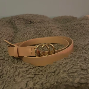 Beige bälte med gulddetaljer - Snyggt beige bälte med dubbla guldringar som spänne. Perfekt för att ge en stilren touch till din outfit. Tillverkat i ett slätt material som ger en elegant look.