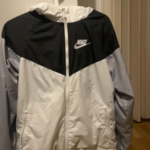  vindjacka  Nike - Säljer en stilren svart och vit vindjacka från Nike. Jackan har en dragkedja framtill och en praktisk huva. Perfekt för blåsiga dagar och har en sportig look med Nike-loggan på bröstet.