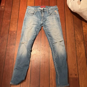Blå jeans från replay - Säljer ett par snygga ljusblå jeans från replay  med slitna detaljer på knäna. De har en klassisk femficksdesign och en knappgylf. Perfekta för en avslappnad stil. 