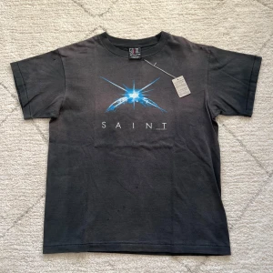 Svart t-shirt med blått tryck från Sonic - Säljer en svart t-shirt från Sonic med ett coolt blått tryck och texten 'SAINT' på framsidan. T-shirten har en klassisk passform och är kortärmad, perfekt för en avslappnad stil. Materialet är mjukt och bekvämt. Made in Japan 