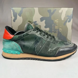 Valentino Rockrunner i camouflage - Snygga Valentino Rockrunner sneakers i ett coolt camouflage-mönster. Skorna har en mix av gröna och svarta nyanser med en röd detalj på hälen. Tillverkade i skinn och mocka med en bekväm sula. Perfekta för att ge din outfit en edgy touch. Endast box medföljer. Ny pris 6599