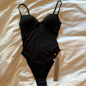 Svart body med justerbara axelband - Snygg svart body med justerbara axelband och vadderade kupor. Materialet är mjukt och stretchigt för en bekväm passform. Helt oanvänd med lappar kvar.